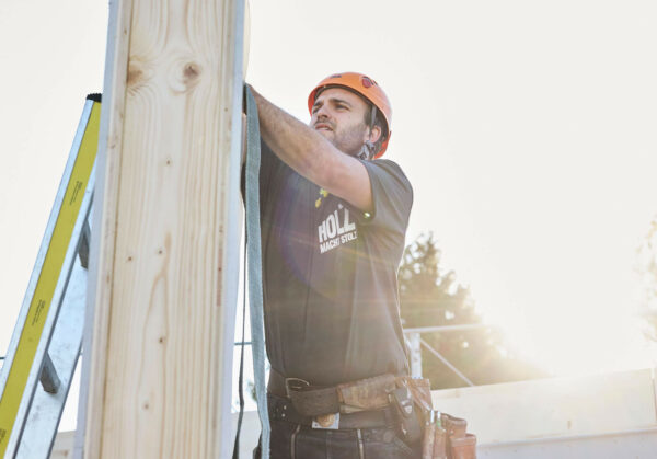Das ist ein Foto. Ein Zimmermann arbeitet auf der Baustelle an einem Holzelement. Der Zimmermann trögt einen orangen Helm und hat eine Werkzeugtasche um die Hüfte gebunden. Gegenüber dem Handwerker ist eine Leiter an das Holzelement angelehnt. Im Hintergrund scheint die aufgehende Sonne hell.