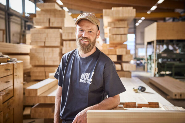 Das ist ein Foto. Es zeigt einen Zimmermann in einer Zimmerei. Der Holzbauer schaut direkt in die Kamera und trägt ein dunkelgraues Holz macht stolz T-Shirt. Das Foto ist ein Symbolbild für Holzbauer, die Gesuche an die Berufsförderung einreichen.
